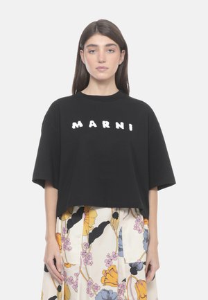 Donna con capelli castani lisci che indossa una t-shirt nera Marni e una gonna floreale con fiori gialli, rosa e blu su sfondo chiaro.