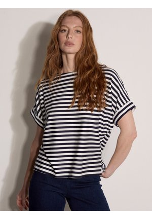 Jeune femme aux longs cheveux roux portant un t-shirt à manches courtes rayé noir et blanc et un jean bleu foncé, debout devant un fond clair uni.