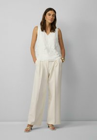 Weiße ärmellose Bluse mit floraler Stickerei, kombiniert mit lockeren cremefarbenen Hosen. Das Model trägt römische Sandalen in Weiß und ein massives goldfarbenes Armband.