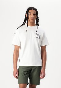 Weißes Baumwoll-T-Shirt mit kurzen Ärmeln, das ein schwarzes Druckdesign auf der linken Brust zeigt. Kombiniert mit olivgrünen Shorts.