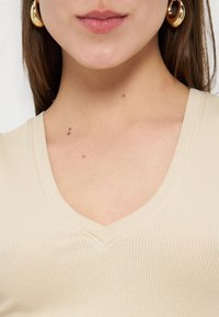 Κοντινή λήψη του λαιμού και του κάτω μέρους του προσώπου μιας γυναίκας που φοράει χρυσούς κρίκους στα αυτιά και μπεζ ριμπ μπλούζα με V-λαιμόκοψη.