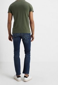 Olivgrünes T-Shirt mit kurzen Ärmeln, kombiniert mit dunkelblauen Slim-Fit-Jeans und weißen Sneakers, zeichnet sich durch ein einfaches Design und minimale Details aus.