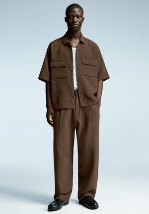 Brown short-sleeve shirt with two chest pockets and matching loose trousers; worn with black shoes. Smooth texture, open collar design.