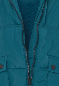 Ein türkisfarbener Jacke mit einem teilweise offenen Reißverschluss, der das dunkelgrüne Fleecefutter zeigt, und mit geknöpften Klappentaschen auf jeder Seite.