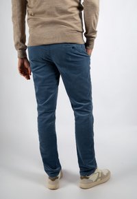 Man staat met handen in de zakken, draagt blauwe corduroy broeken, beige trui en witte sneakers op een witte achtergrond.