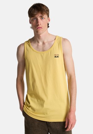 Jonge man met lichte huid en bruin haar, gekleed in een geel mouwloos tanktopje met een klein golflogo op de borst, staand tegen een witte achtergrond.