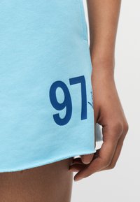 Κοντινό πλάνο ενός χεριού που ακουμπά δίπλα σε μια ανοιχτό μπλε φούστα με τον αριθμό 97 τυπωμένο σε σκούρο μπλε κοντά στο στρίφωμα.