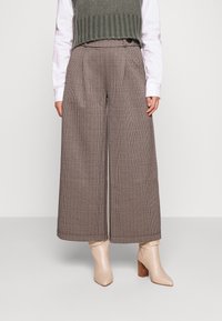 Braune und beige Hahnentrittmuster-Hosen mit weitem Bein, hoch geschnitten mit Falten, kombiniert mit einem grünen gerippten Stricktop und beigen Absatzstiefeln.