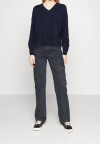Granatowy sweter w serek z ribbowanym wykończeniem, zestawiony z luźnymi, ciemnymi dżinsowymi spodniami cargo z bocznymi kieszeniami i kontrastującym przeszyciem, noszony z czarnymi trampkami.