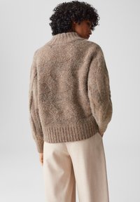 Maglione marrone lavorato a maglia con colletto alto, pattern a texture e orlo a coste. Indossato sopra pantaloni chiari, con una vestibilità morbida.