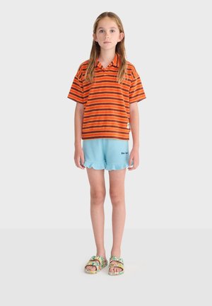 Dieťa s dlhými vlasmi stojí čelom dopredu, oblečené v oranžovo-hnedom pruhovanom polokošeli, svetlo modrých šortkách a kvetinových sandáloch.