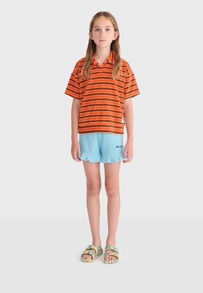 Enfant aux longs cheveux debout face à l'avant, portant un polo rayé orange et marron, un short bleu clair et des sandales fleuries.