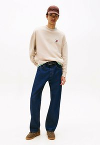 Beige Sweatshirt mit Logo, blaue weit geschnittene Jeans, braune Sneakers und eine braune Cap. Schlichtes Design und legere Kleidung.