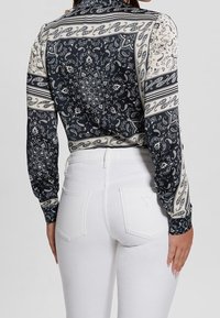 Blouse à manches longues présentant un motif cachemire bleu foncé et crème, avec une silhouette ajustée et un col montant, associée à un jean blanc.