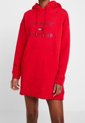 Czerwona, oversize sukienka-bluza z napisem „Tommy Hilfiger” i logo z kryształkami, z przednią kieszenią i kapturem ze sznurkiem, noszona przez osobę.