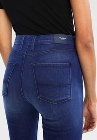 Mörkblå denimjeans med hög midja, bakfickor och en logotyp på midjebandet. Tyget har en lätt blekning och en slät textur.