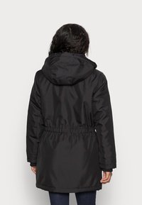 Musta parka, jossa on huppu, kiristettävä vyötärö ja pitkät hihat. Sileä, vettähylkivä kangas. Polven yläpuolelle ulottuva, suora helma.