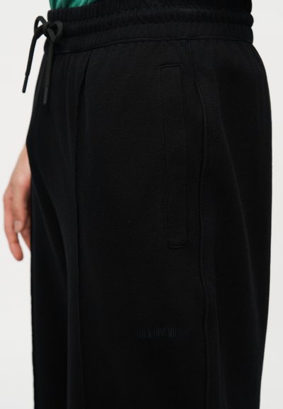 Κοντινό πλάνο μαύρου παντελονιού με κορδόνι και πλαϊνή τσέπη, που φοράει ένα άτομο με πράσινο τοπ.