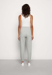 Woman with curly dark hair stands facing a wall wearing a white sleeveless top, gray sweatpants, and white Nike sneakers.