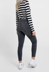 Kobieta w długim rękawie bluzce w czarno-białe paski, ciemnoszarych spodniach z wysokim stanem i białych trampkach za kostkę, widok z tyłu.