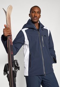 Tengerészkék és fehér sídzseki kapucnival, cipzáras zsebekkel és állítható mandzsettákkal, amelyet egy síléceket fekete kötésekkel tartó személy visel.