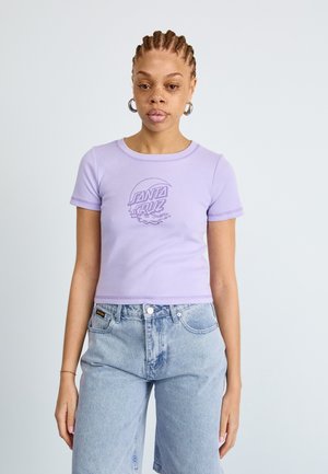 Lillā krāsa kropts t-krekls ar apaļu kaklu, īsām piedurknēm un drukātu "Santa Cruz" logo tumšākā violetā krāsā, kombinēts ar gaiši ziliem džinsu šortiem.
