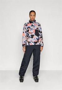 Pull en polaire à motifs abstraits colorés avec une poche noire ; associé à un pantalon sombre ample et des bottes de ski noires.