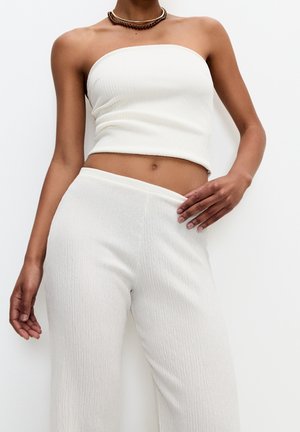 Femme portant un haut court blanc sans bretelles et un pantalon taille haute texturé assorti, accessoirisés avec des colliers superposés.