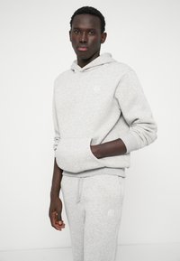 Pilkos medvilnės sportinė džemperis su priekiu kišenėje, raukšlėtais rankogaliais ir mažuoju logotipu. Derinamas su atitinkamais pilkais sportiniais šortais, pabrėžiančiais laisvą siluetą.