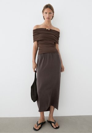 Donna in piedi contro un muro bianco, indossa un top marrone con spalle scoperte, una gonna midi scura, sandali, collana di perle e tiene una borsa nera.