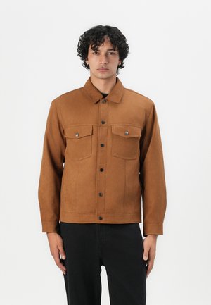 Chaqueta de ante marrón con dos bolsillos en el pecho, botones a presión y cuello con solapa, combinada con pantalones negros. Textura suave y diseño ajustado.