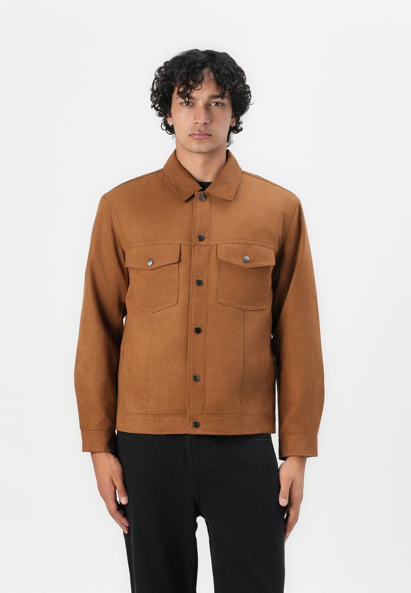 Bruine suede jas met twee borstzakken, drukknoopsluitingen en een kraag, gestyled met zwarte broek. Gladde textuur en strakke pasvorm.