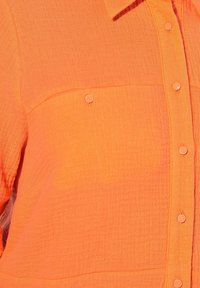 Camicia arancione con bottoni in tessuto testurizzato, dotata di due tasche sul petto e bottoni metallici. Il design è strutturato con un colletto classico.