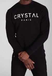 Sweat-shirt noir à manches longues et col rond, avec un grand texte blanc "CRYSTAL PARIS" sur le devant. Tissu doux avec une coupe ajustée.
