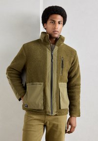 Grüne Fleecejacke mit einem Reißverschluss vorne, hohem Kragen und Aufsetztaschen. Verfügt über einen glatten Stoff an den Ärmeln und einer Reißverschlusstasche auf der Brust.