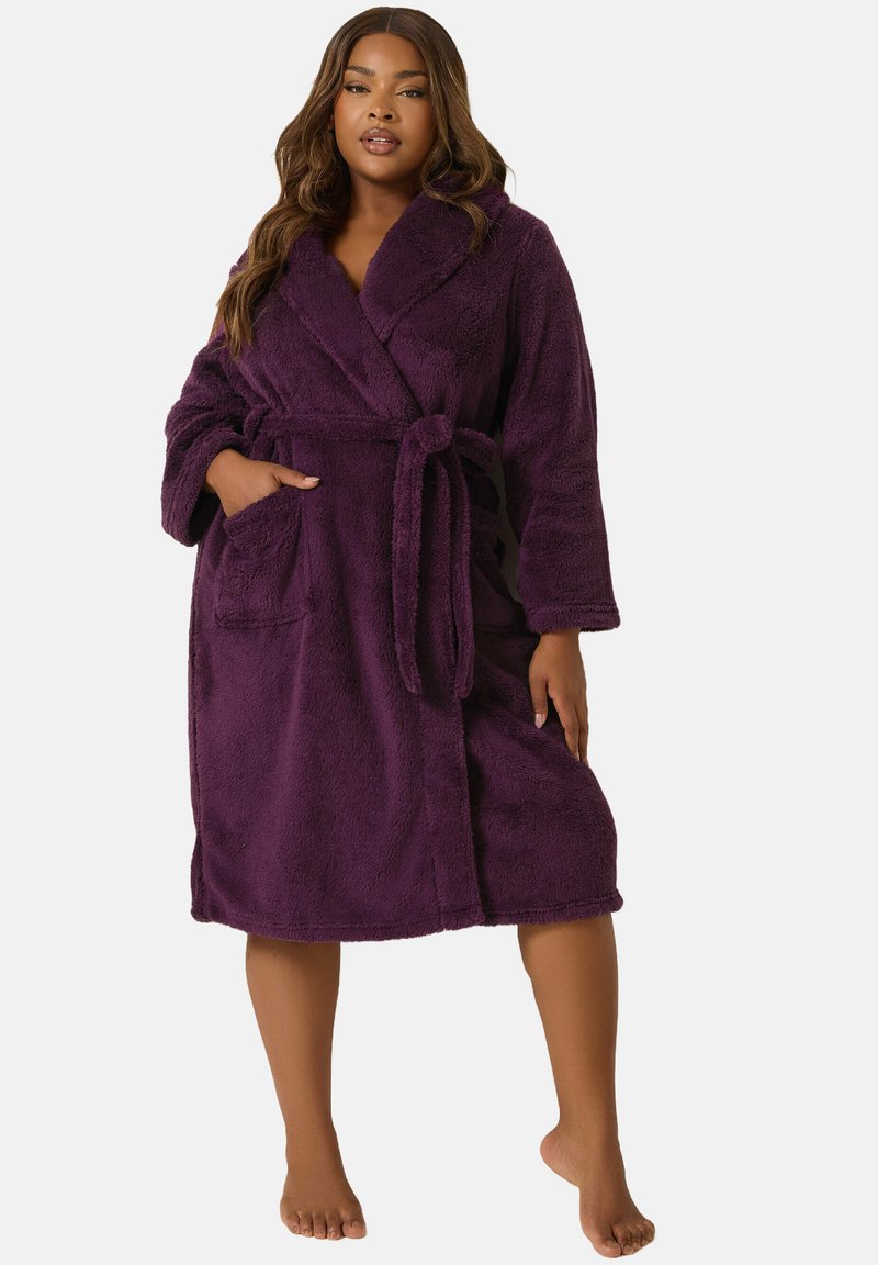 Donna in piedi a piedi nudi, indossa un accappatoio viola di pile di lunghezza al ginocchio con collo a scialle, tasca frontale e cintura legata in vita.