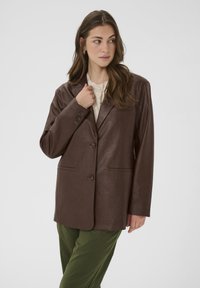 Frau trägt ein braunes Lederblazer über einem beigen Stricktop und grünen Hosen, schaut zur Seite mit einer Hand, die den Revers des Blazers berührt.