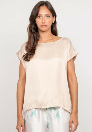 Femme aux longs cheveux bruns portant un haut ample en satin beige à manches courtes et un pantalon blanc avec des motifs floraux bleu clair, debout devant un fond uni.