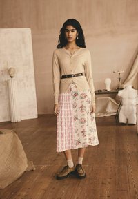 Beige gestrickte Strickjacke mit rundem Halsausschnitt, kombiniert mit einem mehrfarbigen, gefalteten Rock mit floralen und gestreiften Mustern. Braune Plateauschuhe.