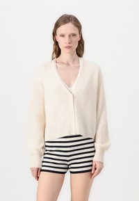 Cremefarvet strikket cardigan med V-hals, frontknapper og lange ærmer; båret over stribede sorte og hvide shorts. Blød tekstur, cropped design.