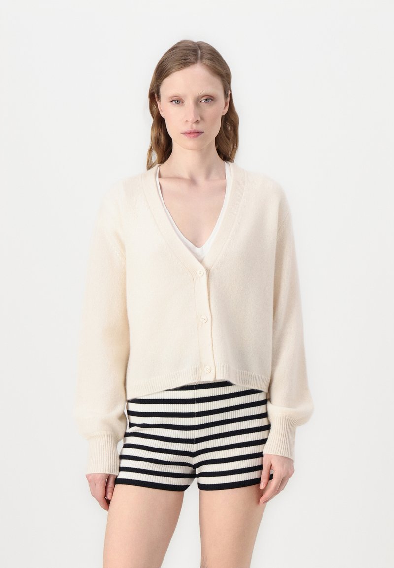 Cremefarvet strikket cardigan med V-hals, frontknapper og lange ærmer; båret over stribede sorte og hvide shorts. Blød tekstur, cropped design.