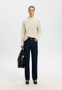 Beiger gerippter Pullover, dunkelblaue hochgeschnittene Jeans, schwarzer Gürtel, schwarze Handtasche und schwarze Ballerinas. Neutraler Hintergrund. Einfacher, lässiger Look.