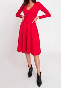 Robe longue rouge à manches longues avec un corsage ajusté, un décolleté en V profond et une jupe évasée, associée à des bottines noires pointues. Tissu à texture lisse.