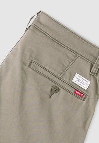 Πίσω τσέπη μπεζ παντελονιού Levi's με κλείσιμο με κουμπί και ραμμένες λευκές και κόκκινες ετικέτες υφάσματος με το λογότυπο Levi's κοντά στην τσέπη.