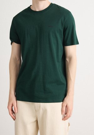 Mann trägt ein dunkelgrünes kurzärmeliges T-Shirt mit Rundhalsausschnitt und hellbeigen Hosen, steht vor einem schlichten Hintergrund.