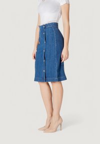 Falda lápiz de mezclilla, azul claro, hasta la rodilla, con botones en el frente, costuras gruesas y un ligero deshilachado en el dobladillo. Se muestran tacones nude a juego.