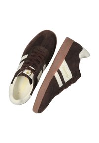 Brązowe zamszowe sneakersy z białymi paskami, teksturowaną gumową podeszwą i logo z boku. Sznurowane pasującymi brązowymi sznurówkami.