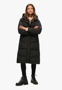 Lang sort pufferjakke med hætte, udstyret med en quiltet design og frontlynlås. Parret med sorte leggings og snørebåndede kampstøvler.