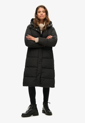 Lange zwarte pufferjas met een capuchon, voorzien van een doorgestikt ontwerp en een ritssluiting aan de voorkant. Gecombineerd met zwarte leggings en veterschoenen.