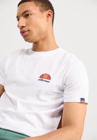 Weiße Baumwoll-T-Shirt mit einem bunten ellesse-Logo auf der Brust und einem kleinen Logo-Detail am Ärmel. Kurze Ärmel, Rundhalsausschnitt.